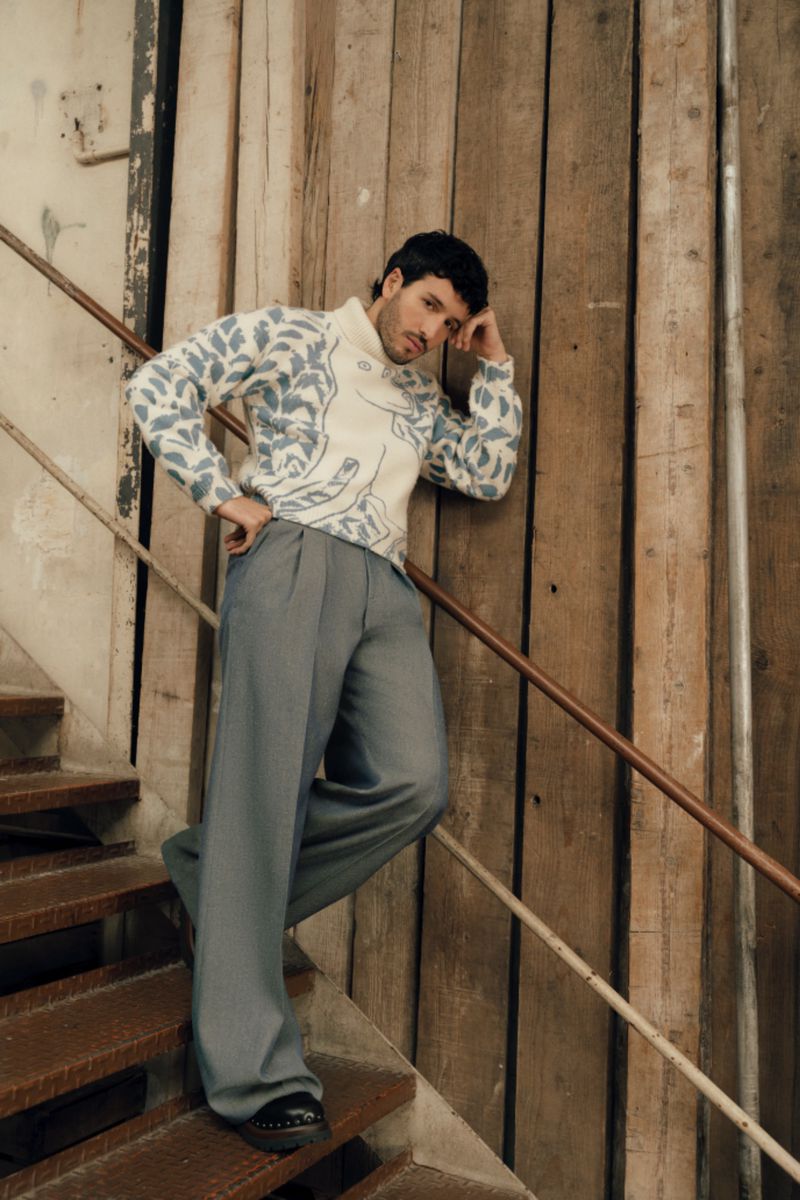 Adult male in a patterned sweater and wide-leg pants poses on indoor stairs with a weathered wood background, captured in a medium shot.