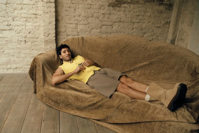 Adult male laying on a tan couch in a photo studio wearing a yellow sweater vest, brown shorts, and boots in a casual pose.