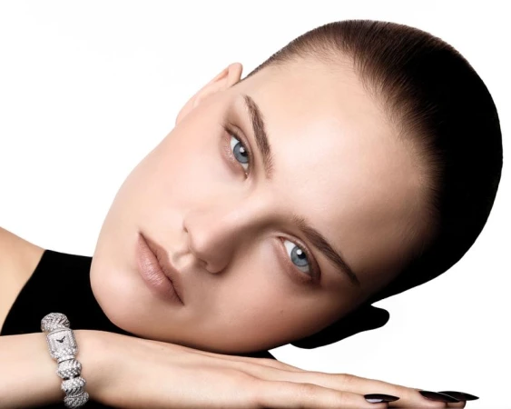 Close-up of a fair-skinned adult woman with blue eyes and short brown hair, posing with a diamond-studded watch on her wrist against white.