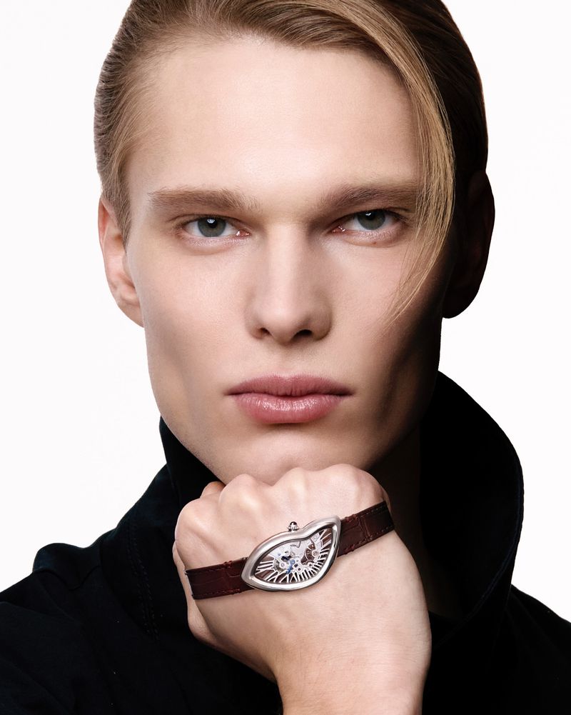 Close-up of a blond male model wearing a luxury wristwatch with a brown strap against a stark white background in a studio setting.