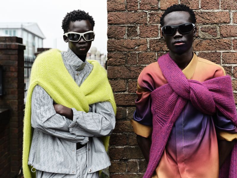 Two dark-skinned men pose by a brick wall outdoors, wearing sunglasses and draped with knit scarves in a fashion-forward outdoor shot.