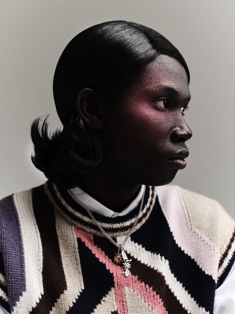 Close-up of a dark-skinned person with side-parted hair, wearing a colorful knit sweater vest and a silver chain necklace in a studio setting.