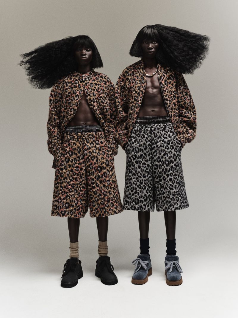 Two dark-skinned individuals with large afros pose in matching animal-print outfits with bare chests against a neutral backdrop, showcasing avant-garde fashion.