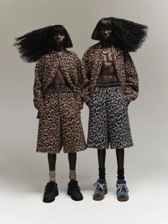 Two dark-skinned individuals with large afros pose in matching animal-print outfits with bare chests against a neutral backdrop, showcasing avant-garde fashion.