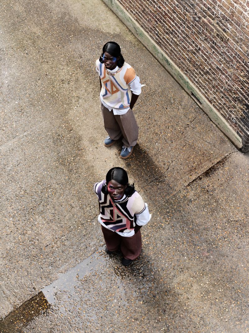 Two black adults wear patterned sweaters and trousers. They stand on concrete in an eye-level shot. A brick wall appears in the background.