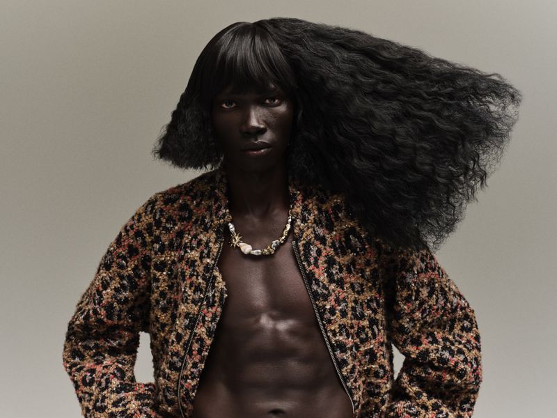 Dark-skinned model with voluminous black hair wears an open leopard print jacket and seashell necklace in a studio portrait.