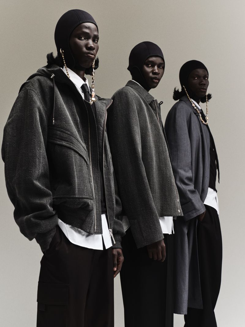 Three Black fashion models in gray coats, white shirts, and dark pants pose in a studio setting. Long gold earrings add a touch of elegance.