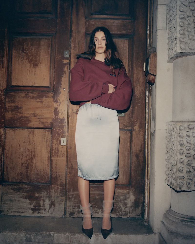 A woman in a red hooded sweatshirt and cream skirt poses in front of a large wooden door with stone architecture in the background.