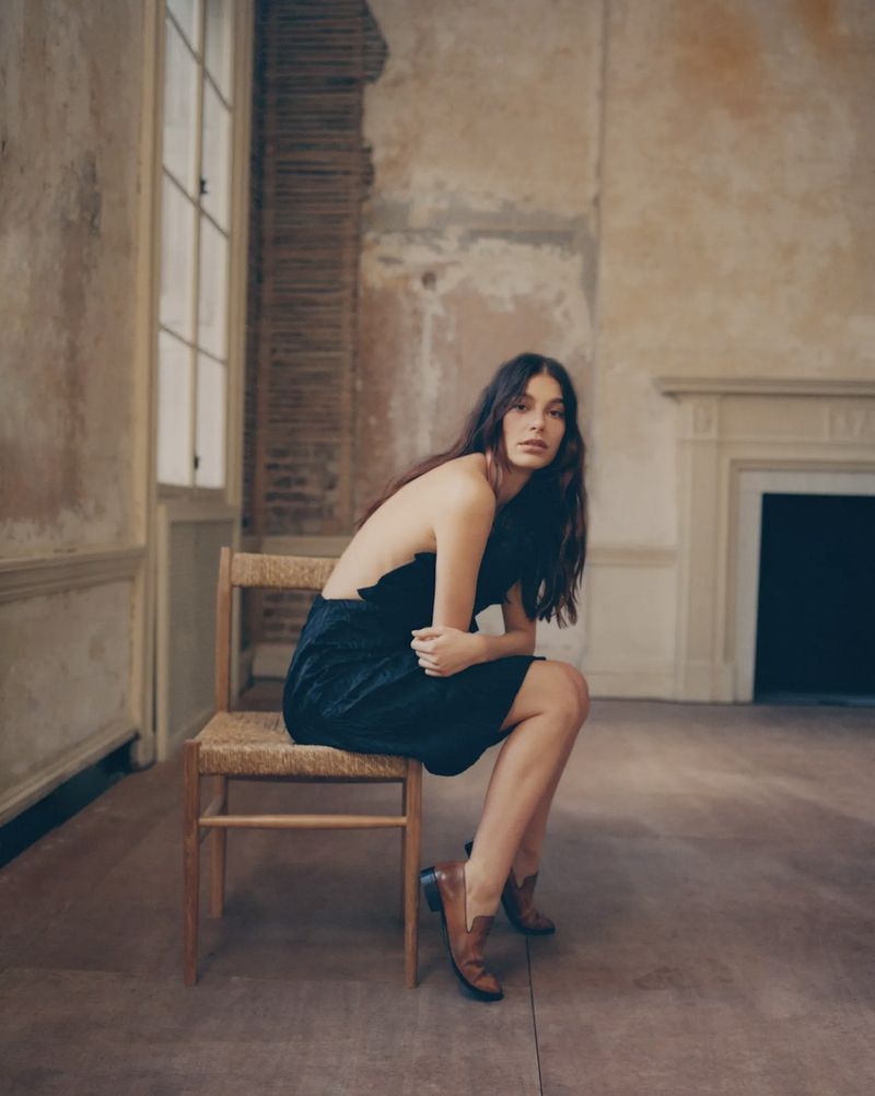 Brunette woman in a black dress and brown boots sits on a wooden chair in a sparse room with an unfinished fireplace.