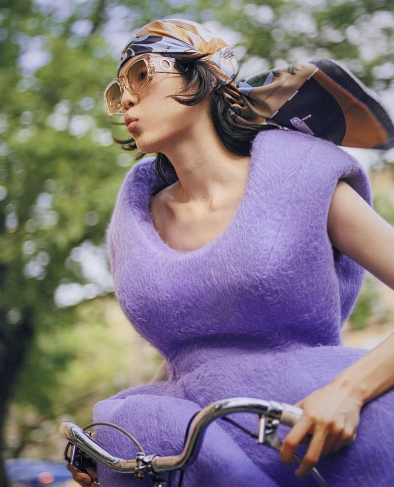 Woman in a purple dress and scarf riding a bicycle outdoors, wearing sunglasses, with green trees in the background on a sunny day.