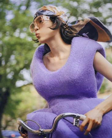 Woman in a purple dress and scarf riding a bicycle outdoors, wearing sunglasses, with green trees in the background on a sunny day.