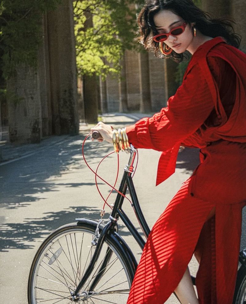A woman in a red outfit rides a black bicycle on a paved road lined with tall trees on a sunny day, showcasing fashion and outdoor lifestyle.