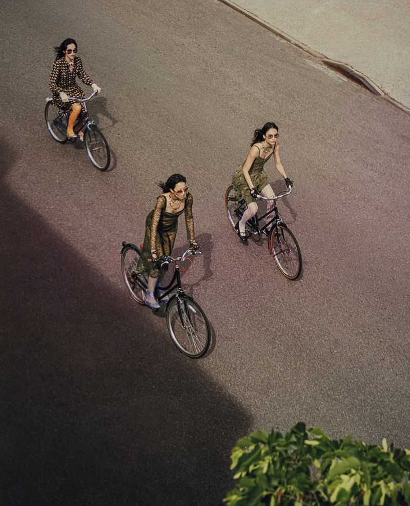 Three women in vintage dresses ride bicycles on an asphalt road, photographed from above with natural light and shadows creating contrast.