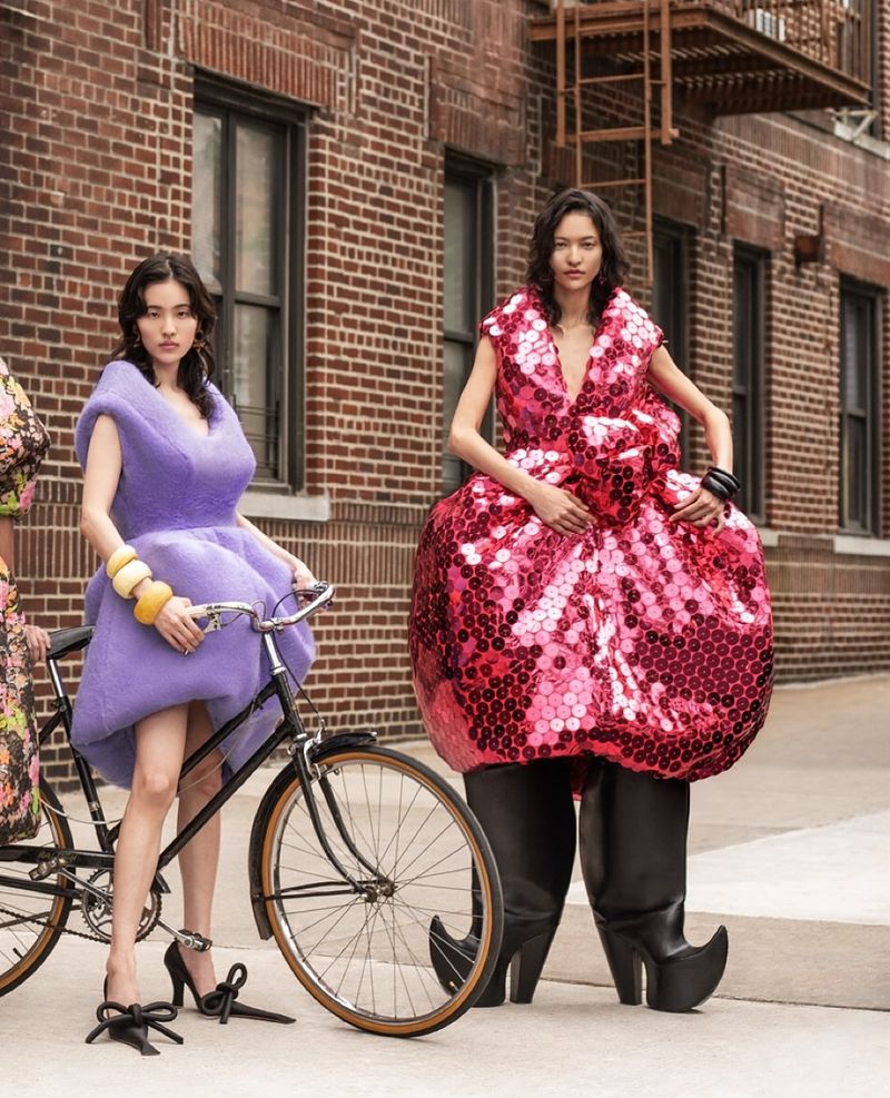 Two fashion models pose outdoors in puffy dresses near bicycles. One is lavender, the other bright pink. A brick wall is in the background.