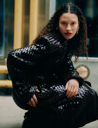 Adult female with curly hair wearing a shiny, textured black coat, crouching on a city street with yellow bars in the background.