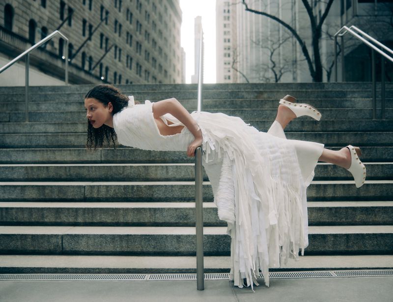 Young adult female model in white fringe dress poses on staircase with hand on rail, wearing white clogs in an urban environment.