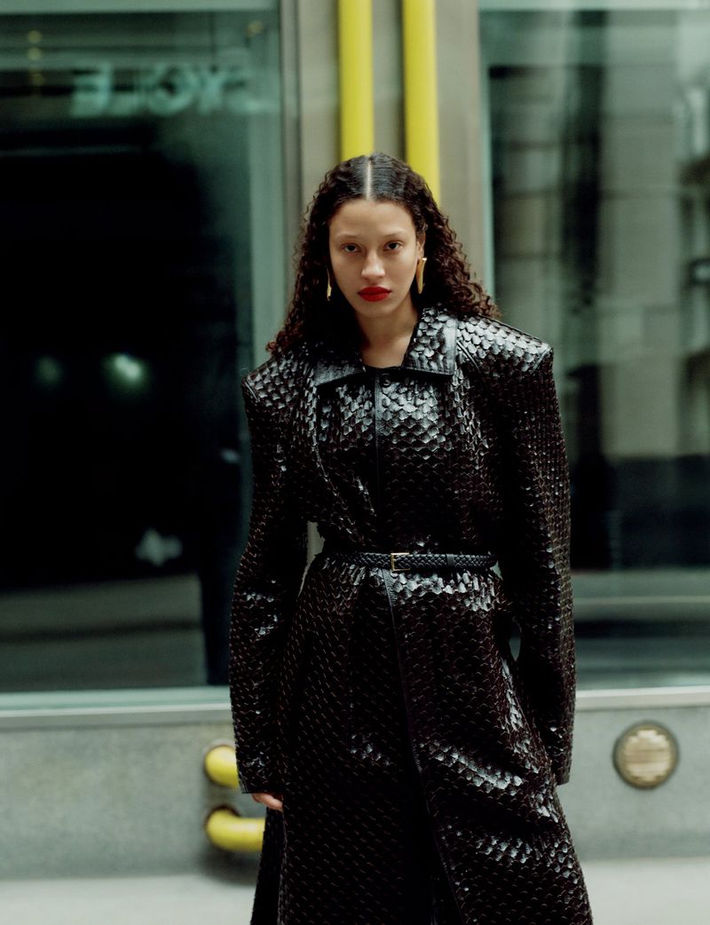 Fashionable woman in a black textured coat with a belt, red lipstick, and gold earrings posing in an urban setting with muted lighting.