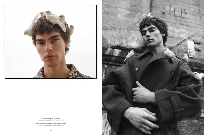 Monochrome fashion shot: a fair-skinned male model wears a coat, perched with white birds against a worn, graffitied wall, edgy style.
