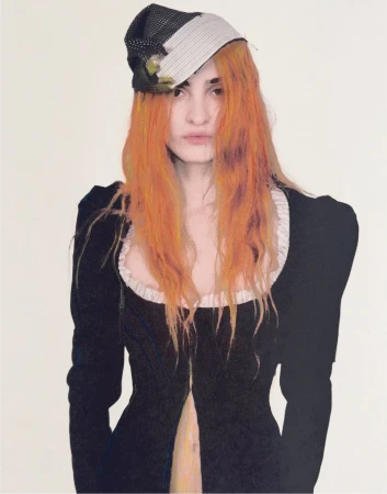 Portrait of a woman with orange hair wearing a black dress with a white collar and a patterned hat against a white background.
