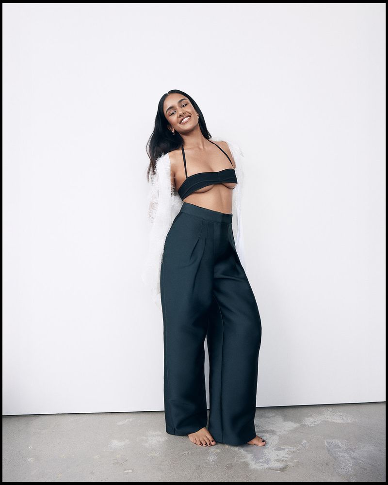 A dark-haired woman in a black bikini top and wide-legged pants poses in front of a plain white wall, looking at the camera and smiling.