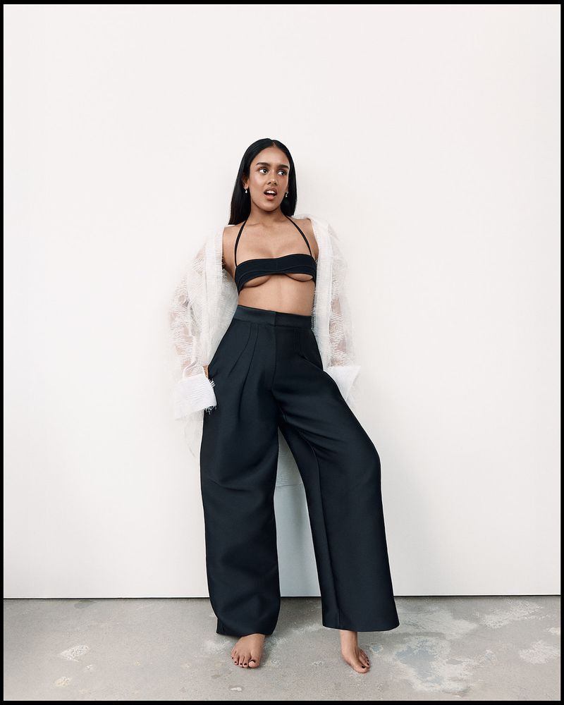 Fashion portrait of an adult woman in a black bikini top, wide black pants, and a sheer white jacket, posing against a white wall.