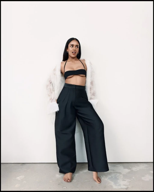 Fashion portrait of an adult woman in a black bikini top, wide black pants, and a sheer white jacket, posing against a white wall.