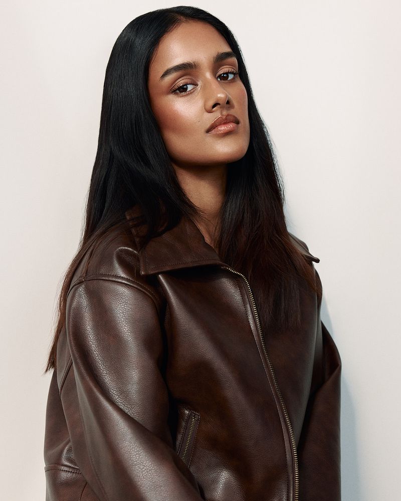 Portrait of a woman with long dark hair wearing a brown leather jacket against a white background in a studio setting.