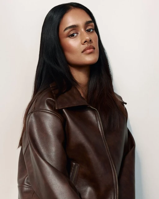Portrait of a woman with long dark hair wearing a brown leather jacket against a white background in a studio setting.