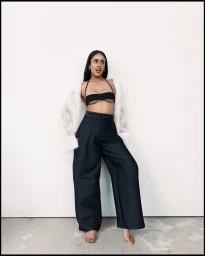 Shabana Azeez barefoot in black wide-leg trousers, a black halter top, and a sheer white coat against a white studio wall.