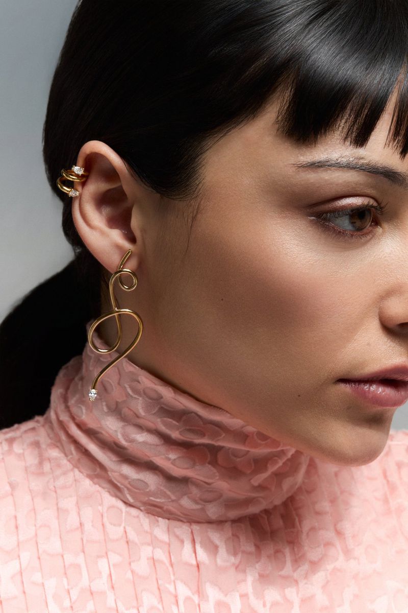 Close-up of a woman with black hair wearing a pink patterned high-neck shirt, a gold spiral earring and a double-ring earring.