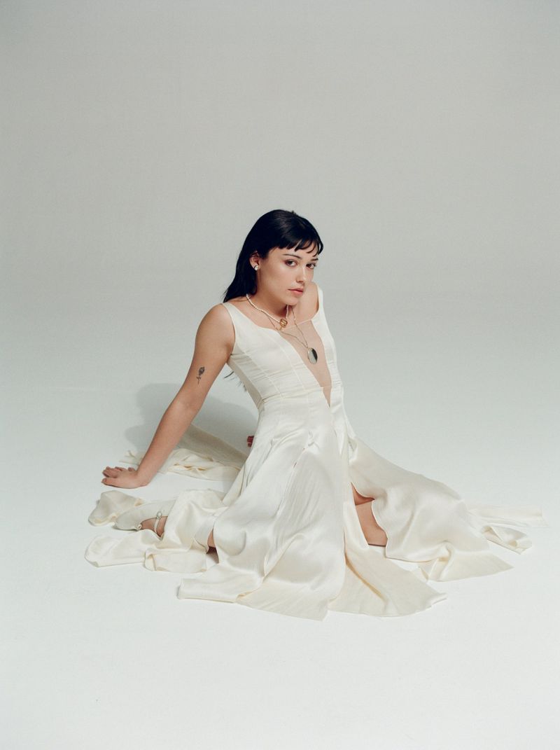 Brunette woman in a white sleeveless dress sits on the floor in a light studio, wearing jewelry, with a neutral facial expression.