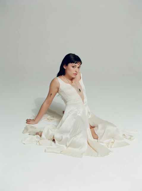 Brunette woman in a white sleeveless dress sits on the floor in a light studio, wearing jewelry, with a neutral facial expression.