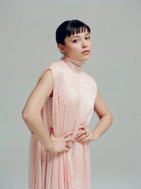 Portrait of a young adult woman with short black hair wearing a light pink dress with hands on hips against a neutral background.