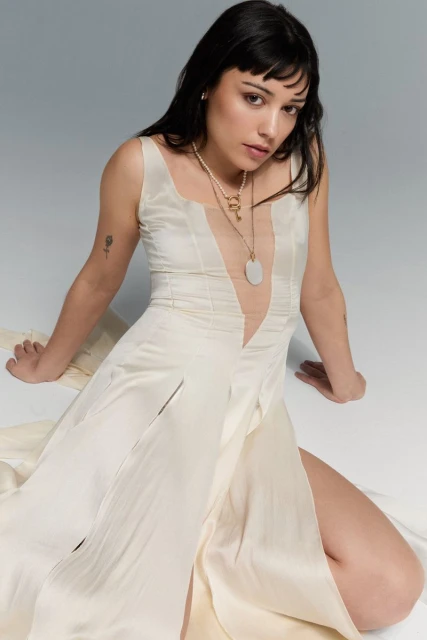 Young woman in an off-white dress with a thigh-high slit poses in a studio with a gray background. She wears necklaces and has dark hair.