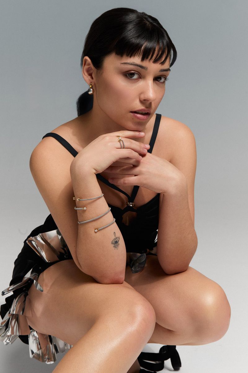 Woman in black dress poses in a photo studio setting with silver accents. She wears jewelry and has an arm tattoo.