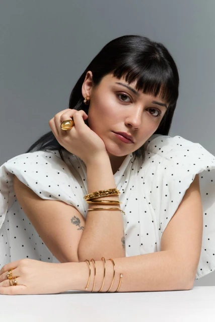 Close-up studio portrait of a young adult woman wearing gold jewelry and a white polka dot shirt, looking at the camera.