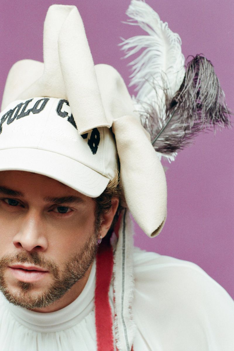 Close-up of a fair-skinned man with a beard wearing a white Polo cap adorned with feathers, against a plain purple backdrop.