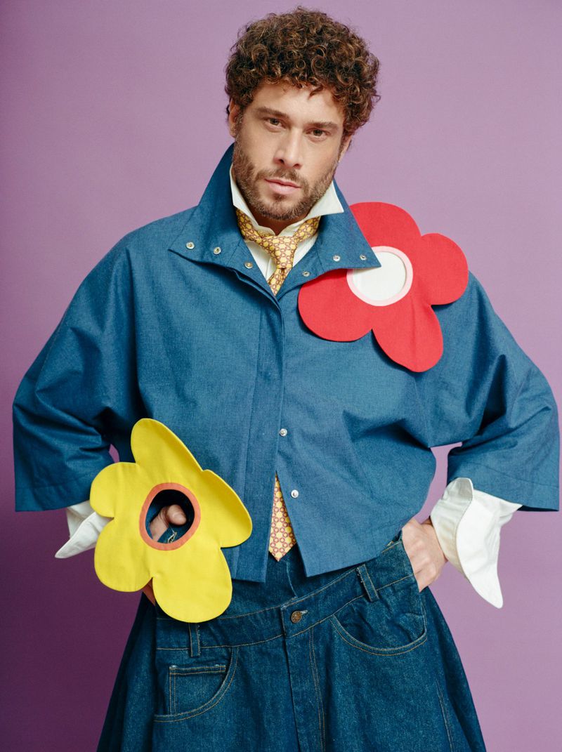 Adult male fashion model poses in denim jacket and jeans skirt with handmade yellow and red flower adornments on a purple background.