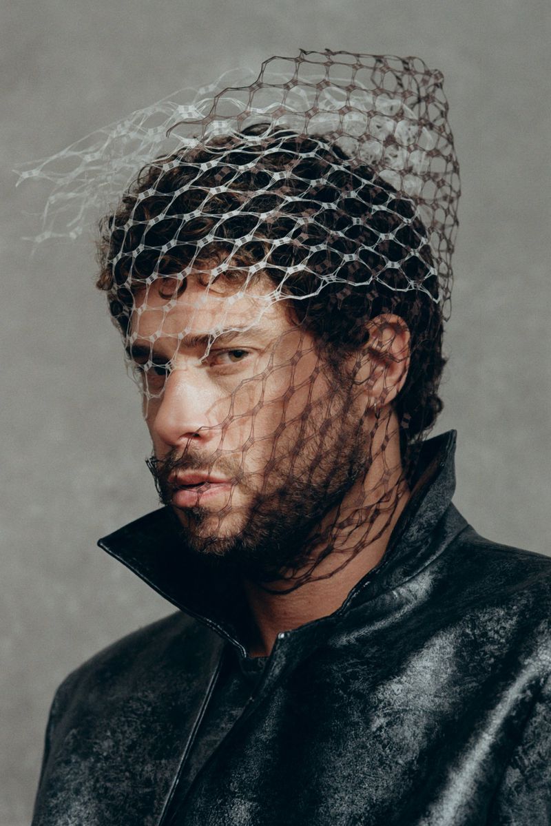 Headshot of an adult male with curly hair and a beard, wearing a black leather jacket and a translucent netted head covering.