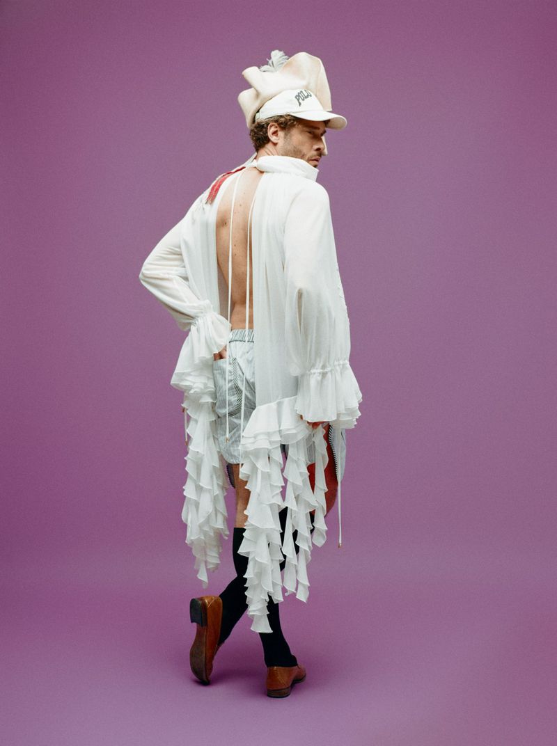 Man posing in white ruffle top and shorts with baseball cap in a studio setting against a purple background, fashion editorial shot.