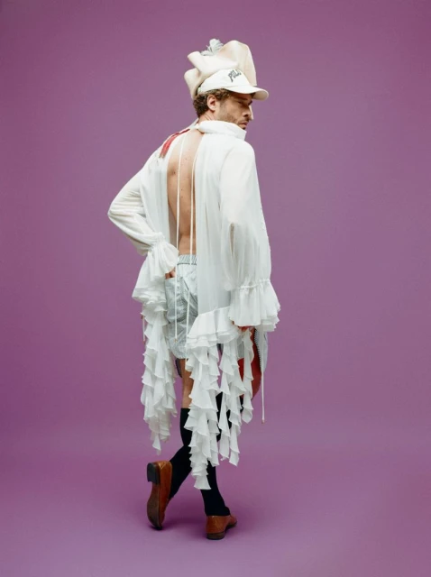 Man posing in white ruffle top and shorts with baseball cap in a studio setting against a purple background, fashion editorial shot.