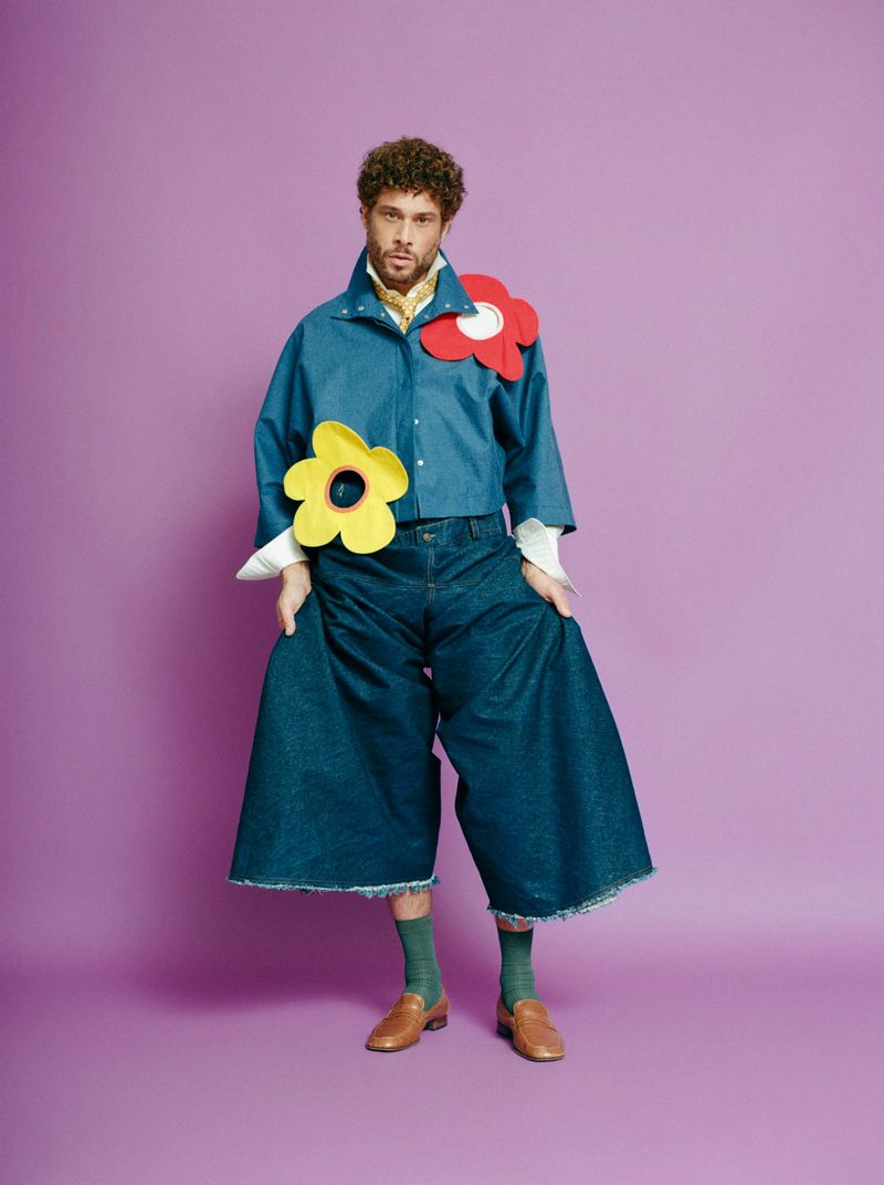 Man in oversized denim outfit with flower accents posing against a purple backdrop in a fashion forward editorial style.