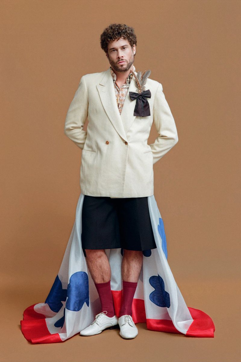 Full-shot of a man with curly brown hair in an off-white double-breasted blazer and black shorts, standing on a tan backdrop with patterned fabric.
