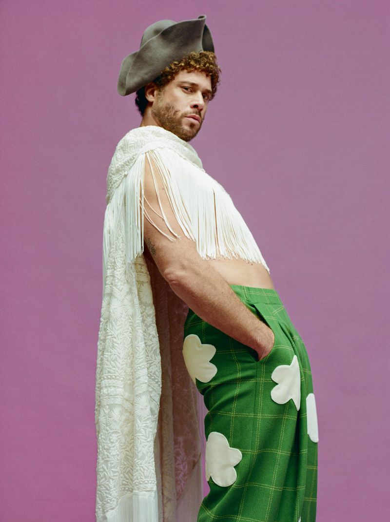 Man in gray hat, white shawl with fringe, and green pants with white flowers, posing against a solid purple backdrop in a studio shot.