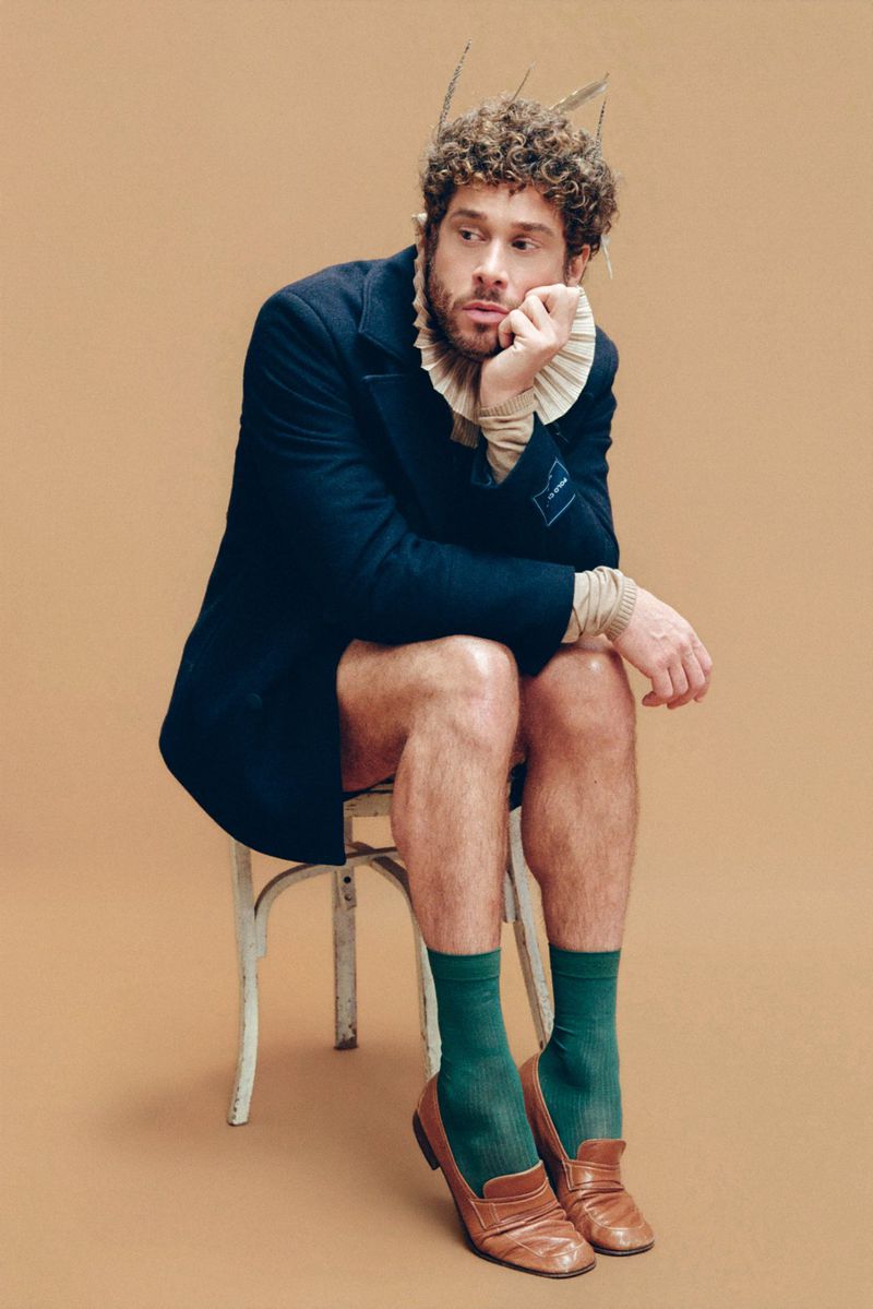 Man in blazer, ruff, and green socks sits on a chair against a peach background in a studio setting, gazing wistfully to the right.