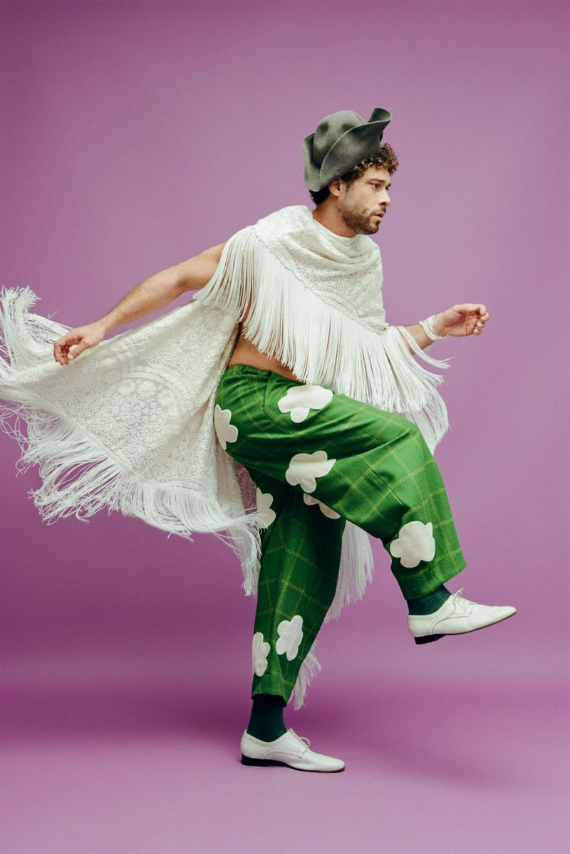 A man in green pants with cloud patterns and a fringed shawl strikes a pose on a purple background, showcasing a unique fashion style.