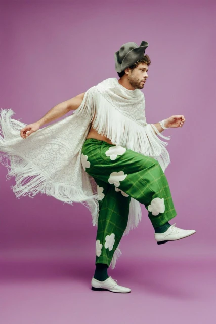 A man in green pants with cloud patterns and a fringed shawl strikes a pose on a purple background, showcasing a unique fashion style.