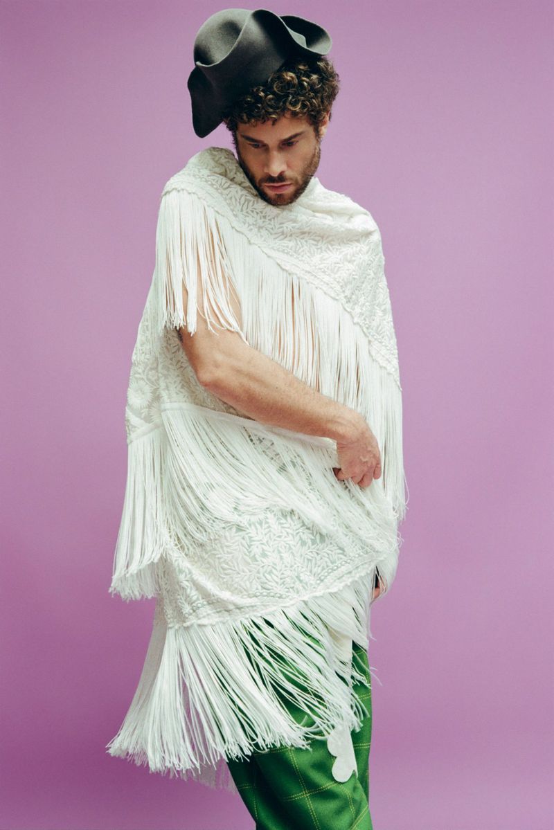 A fashionable person with curly hair poses in a fringed white shawl, wide-brimmed hat, and green pants against a solid purple backdrop.