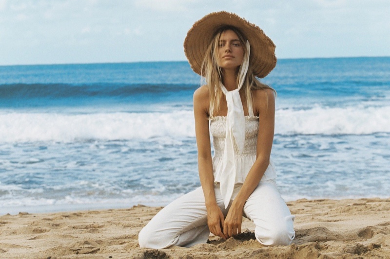 A woman in a white outfit and straw hat poses on a sandy beach with ocean waves in the background on a sunny day.