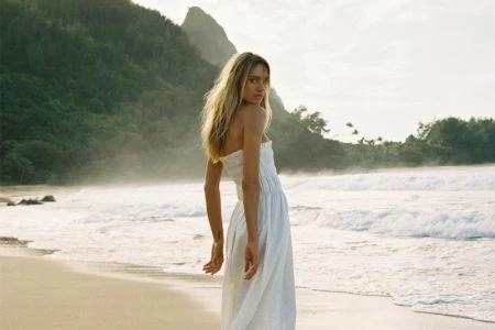 Model wearing a white strapless dress on a sandy beach with breaking waves and a lush green backdrop.